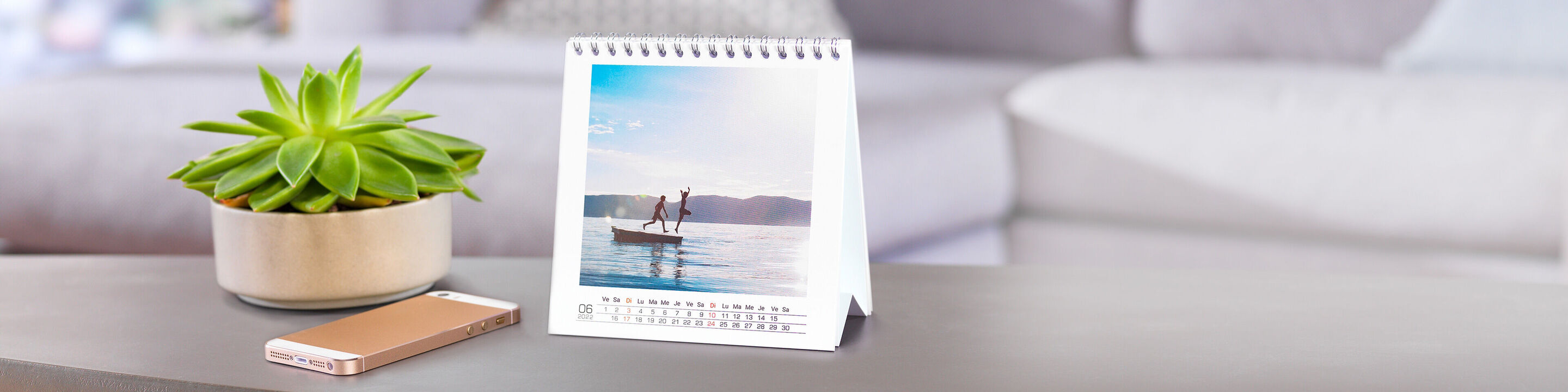 Un calendrier CEWE est sur une table. Une personne est debout dans l'eau sur un paddleboard. Nature paisible en arrière-plan.
