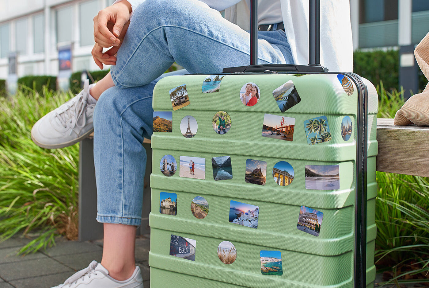 Valise rigide verte décorée de stickers photo de voyage, posée près d’une personne assise sur un banc