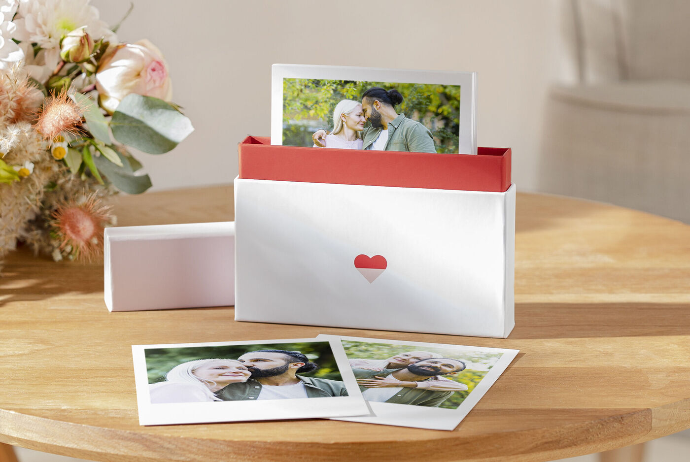Une jolie boîte blanche avec un design en cœur rouge, contenant des photos. La boîte est sur une table en bois, avec quelques photos d'une famille heureuse à côté. De belles fleurs fleurissent en arrière-plan.