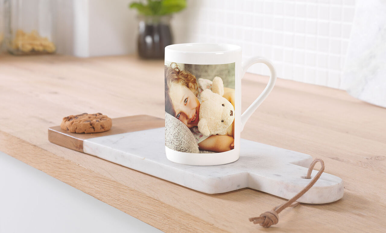 Un enfant étreint une peluche sur une tasse à café. Un biscuit est posé sur une planche en bois.