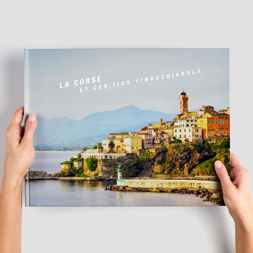 Des mains tiennent un livre photo CEWE avec une image de la côte italienne de Corse.