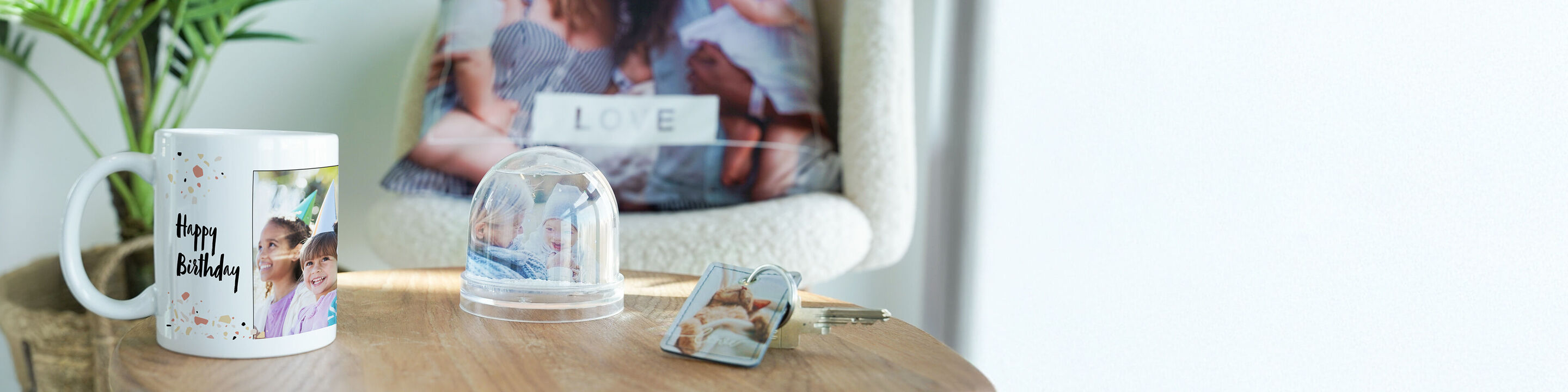Une tasse avec une photo charmante et l'inscription 'Joyeux Anniversaire', une boule en verre avec une photo, un cadre photo sur une table avec un coussin au fond affichant le mot 'Amour'